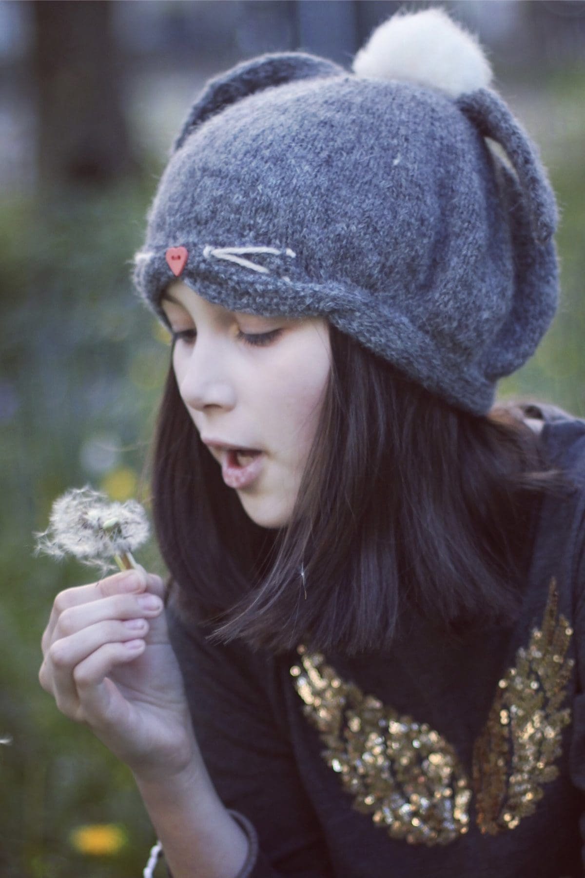 Girl blowing dandelion seeds. Teen with beanie hat making a wish. Springtime fun.