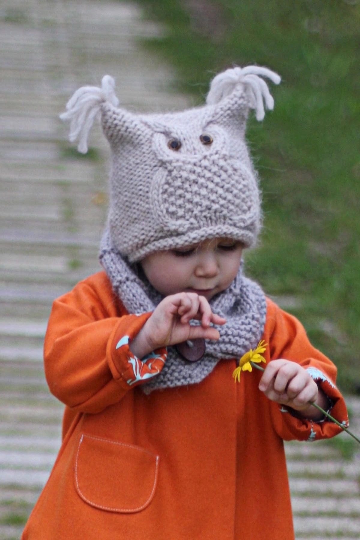 Cute baby girl in knit hat and scarf holds yellow flower. Autumn fashion.