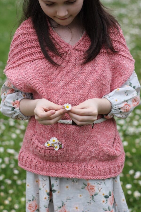 Girl in pink knit dress holds daisy. Floral sleeves, daisy pocket detail, and braided belt. Spring fashion.