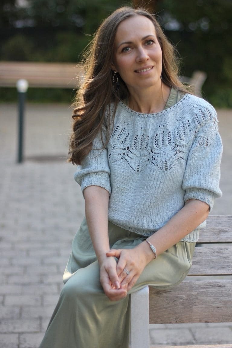 Woman in light blue knit sweater and green skirt sitting on a bench.